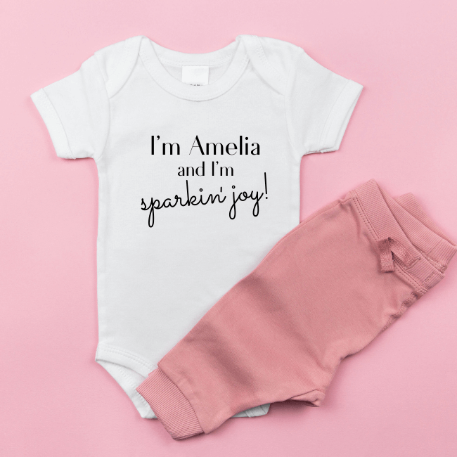 White-baby-onesie-with-transfer-saying-I'm-Amelia-I-m-sparkin'-joy! On a pink background with leggings.